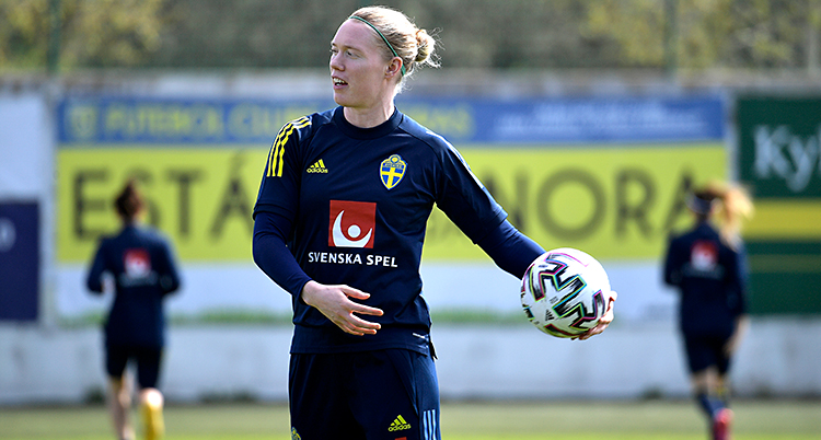Hon står på en fotbollsplan. Hon har träningskläder. Hon har en boll i handen som hon är på väg att kasta.