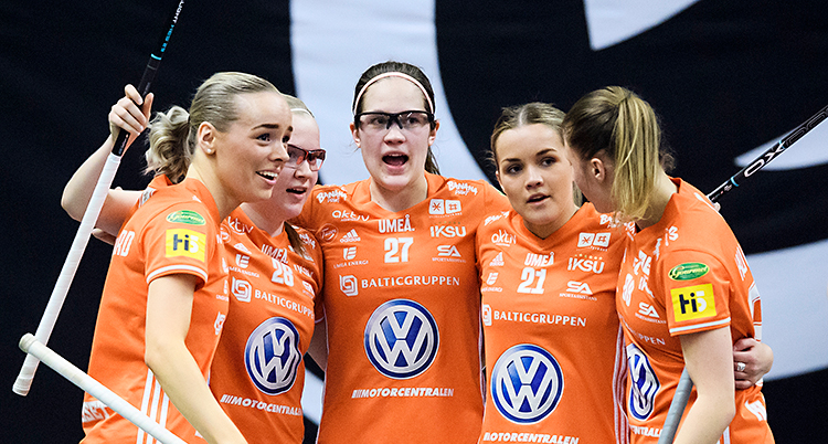 Bilden är från en match i innebandy. Några kvinnor kramas efter ett mål. De har orange tröjor och innebandyklubbar i händerna.