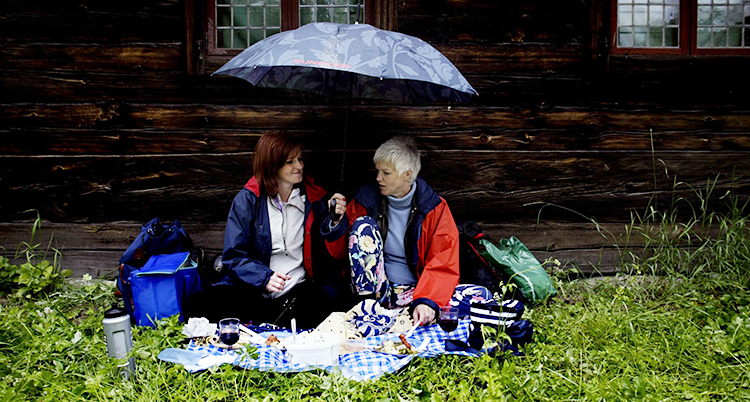 Två personer sitter mot en husvägg. De sitter på en filt. De har picknick. En av dem håller i ett paraply.