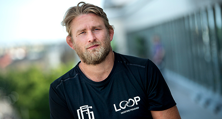 Han tittar in i kameran. Han har blont hår och skägg och en svart t-shirt.