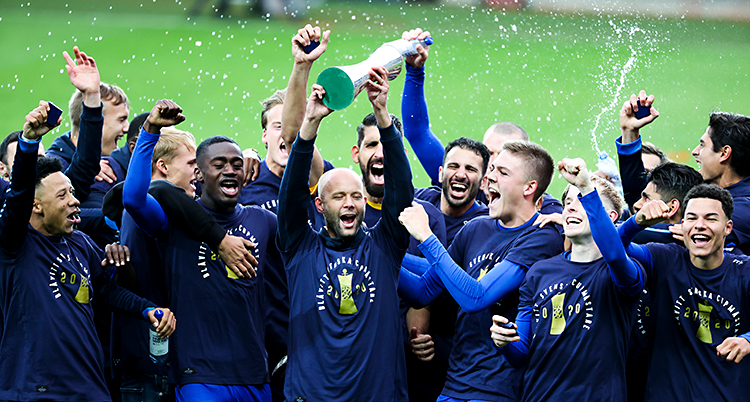 En massa män står på en fotbollsplan. De är glada. De hoppar och jublar. En av dem håller i en pokal.