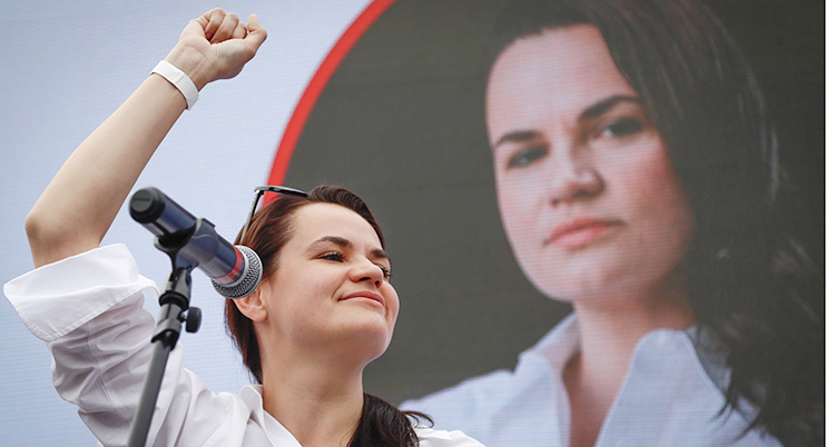 Svetlana Tichanovskaja talar på ett valmöte. Hon har på sig en vit blus och höjer ena handen.