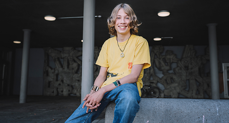 En kille sitter och ler och tittar in i kameran. Han har på sig en gul t-shirt och blå jeans.