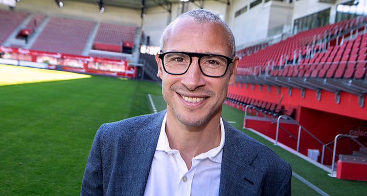 Henrik Larsson ler mot kameran. Han står på en fotbollsarena. Han har kort hår och glasögon. Han har vit skjorta och kavaj.