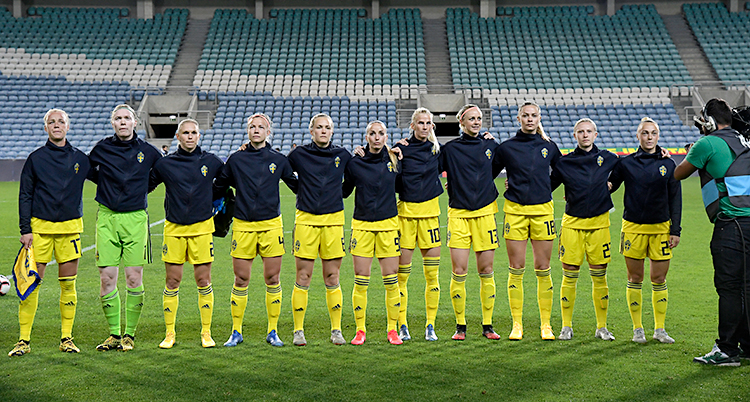 Det svenska laget står bredvid varandra. De är på en fotbollsplan. Snart ska en match börja. De har blå tröjor och gula shorts.