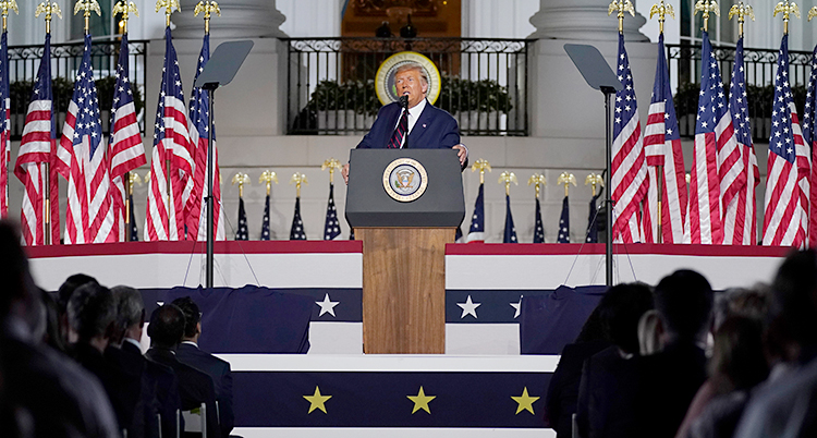 Donald Trump står i en talarstol. Han pratar i en mikrofon. Bredvid honom finns flera amerikanska flaggor. I bakgrunden syns Vita huset.