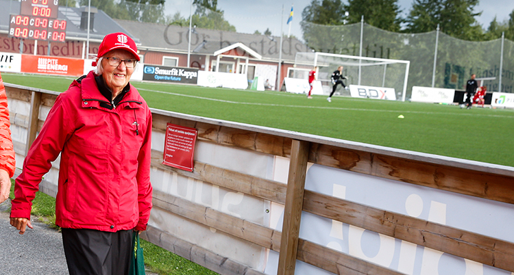 Berit i röd jacka och keps ler och går bredvid fotbollsplanen.