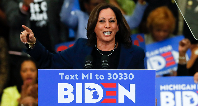 Hon står i en talarstol och ser glad ut. Framför henne är en skyld där det står Biden.