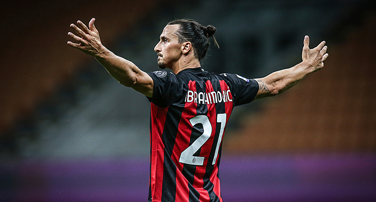 Bilden är från en match i fotboll. Zlatan firar ett mål. Han sträcker ut armarna. Han har håret i en tofs. Hans tröja är randig i svart och rött.