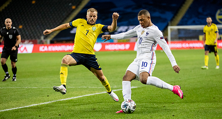 Vi ser en match i fotboll. En fransk spelare har bollen. En svensk spelare försöker ta bollen.
