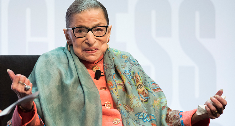Ruth Bader Ginsburg ler mot kameran och slår ut med händerna. Hon sitter i en stol och har en ljusgrön schal runt axlarna.