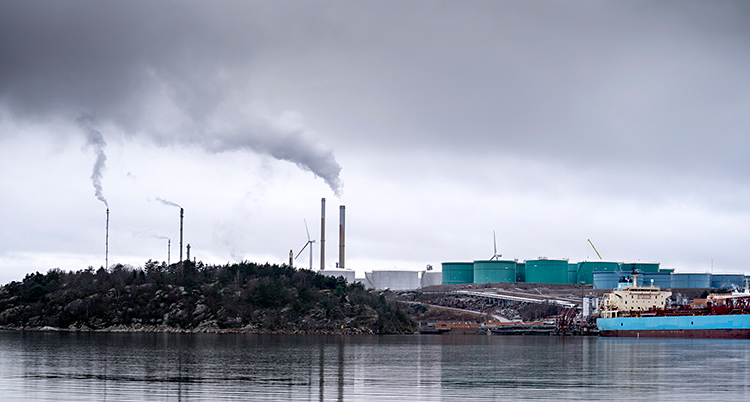 Bilden är tagen från vattnet. Vi ser en stor fabrik. Det kommer rök ur höga skorstenar. En stor båt finns vid fabriken.
