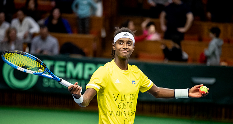 Mikael Ymer har gul tröja och ett vitt hårband. Han ler och håller ett racket i ena handen.