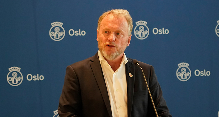 Han har vit skjora och kavaj. Han talar i en mikrofon. Bakom honom står det Oslo på en vägg.