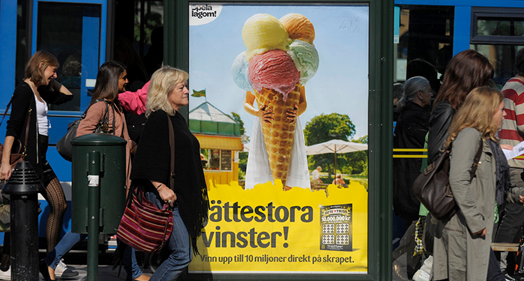 En kvinna går förbi en busshållsplats. Där sitter ne tor skylt med reklam för lotter. Jättestora vinster. Står det på skylten.