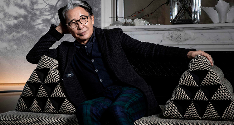Kenzo sitter lutad mot en soffa. Kudden han lutar sig mot ser ut som en pyramid. Han har en svart cape på axlarna och ler mot kameran.