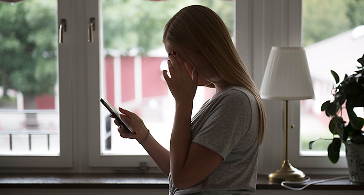 Bilden är tagen inuti ett hus. Vi ser en blond tjej i profil. Hon håller i en mobil och tittar på den. Hon håller upp sin andra hand vid ansiktet, så vi ser inte ansiktet.