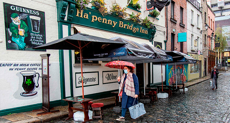 En person går med ett paraply på en regntung gata på Irland. Personen har munskydd och ett paraply över huvudet. I bakgrunden en irländsk pub.