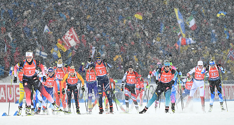 Flera åkare som åker skidor på en stadion. De har gevär på ryggen. I bakgrunden finns en massa publik. Det snöar.