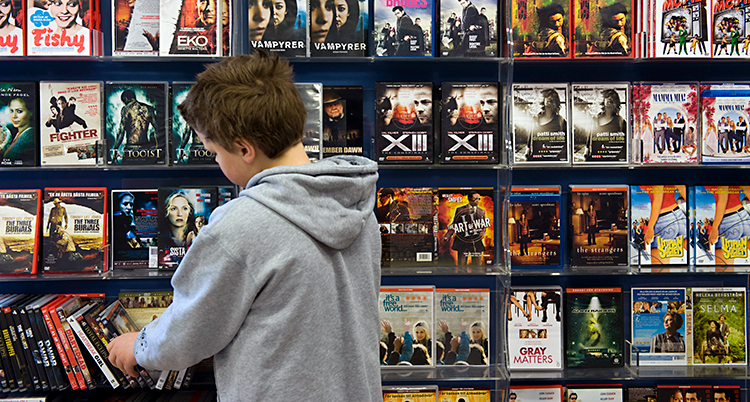 En pojke står och tittar vid en hylla. På hyllan finns det en massa olika dvd-filmer.