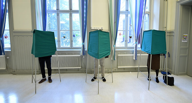tre personer står bakom tre röstbås i en sal. Vi ser bara deras ben.