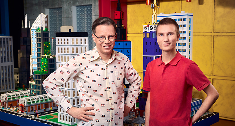 De står bredvid varandra. Den ena har vit skjorta. Den andra röd t-shirt. Bakom dem syns ett stort bygge av lego.
