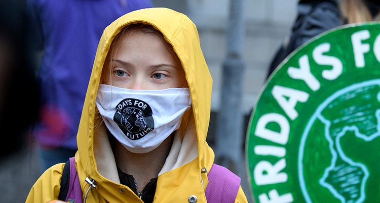 Greta står med gul regnjacka. Hon har munskydd. På det står Fridays for future.