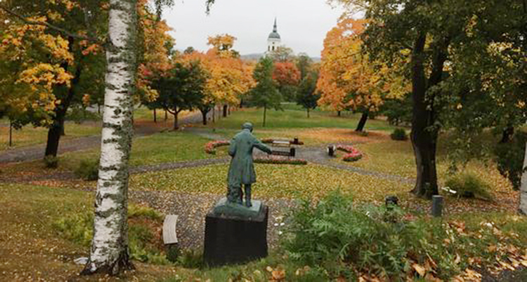 En staty står mitt i en park. Träden har olika färger i grönt och orange och gult.