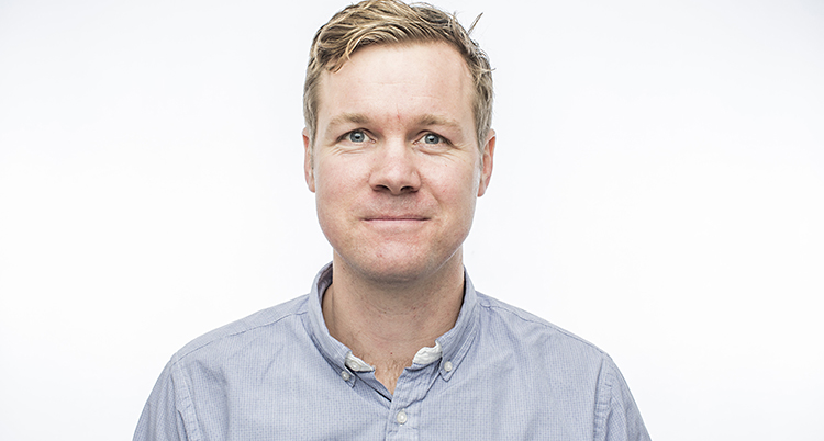 Han tittar in i kameran. Han har blont hår och ljust blå skjorta.