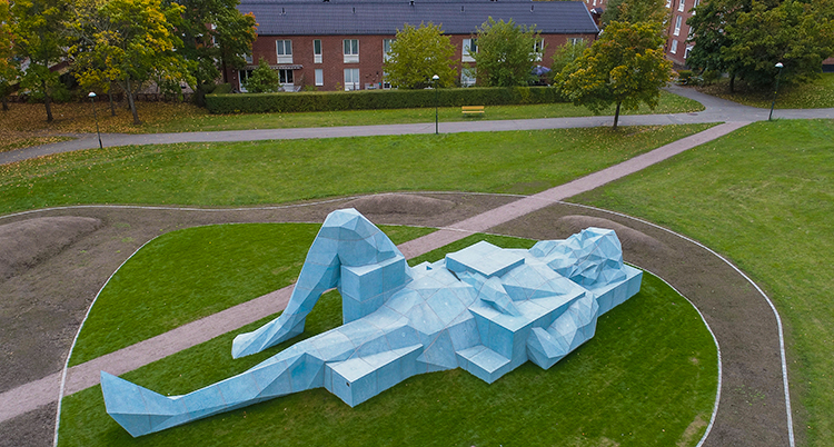 En skulptur i form av en lång, ljusblå person av betong ligger utsträckt i grönt gräs