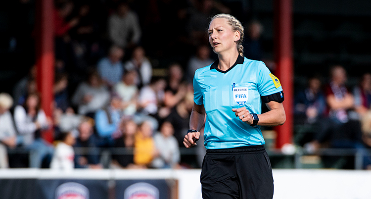 En kvinna med blont hår, klädd i ljusblå domartröja springer på en fotbollsplan med läktare i bakgrunden.