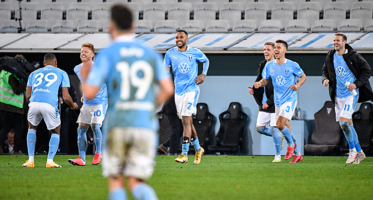 Bilden är från en stadion för fotboll. Spelare i Malmö FF springer på gräset. De är glada. De har ljusblå tröjor.