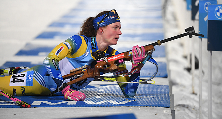 Hanna Öberg lägger sig ned för att skjuta. Hon har fram sitt gevär. Hon har skidor på sig och hon har Sveriges dräkt i gult och blått.