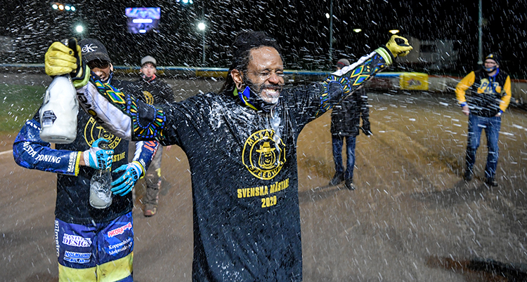 En man med svart hy och svart hår sträcker ut armarna och ler. Han håller i en flaska. Han är klädd i en svart speedwaytröja. Bakom honom står tre män utspridda på en speedwaybana