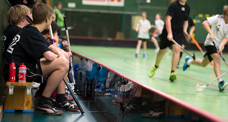 Några pojkar sitter på en bänk bredvid en plan där andra barn spelar innebandy. Pojkarna är avbytare