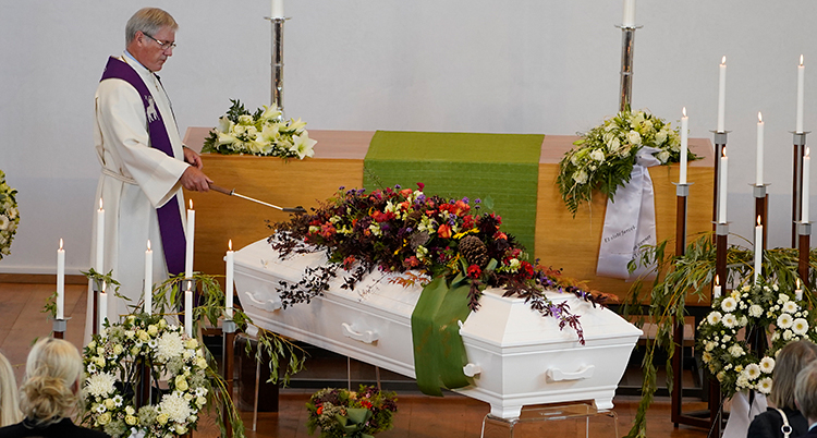 En präst står vid en kista. Kistan har blommor på sig.