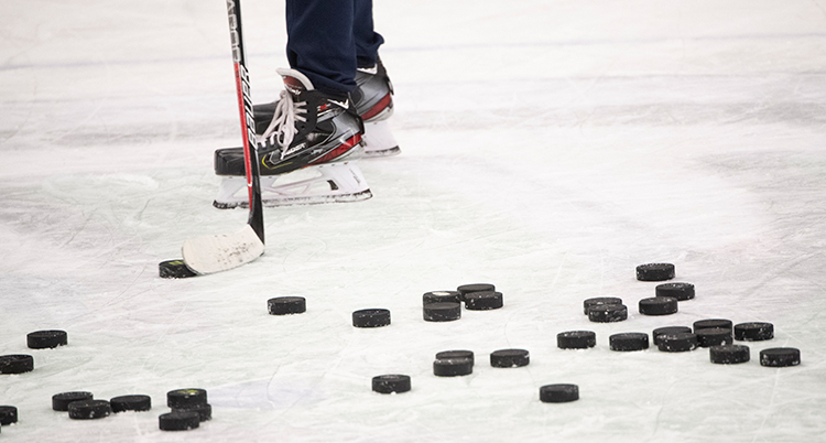 En massa ishockeypuckar ligger på isen och en person med skridskor och ishockeyklubba står bakom puckarna