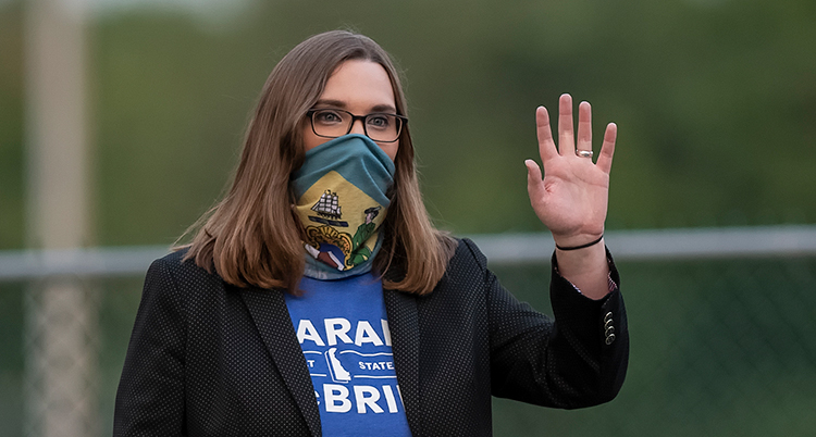 Hon vinkar på bilden. Hon har långt brunt hår och glasögon. För munnen har hon ett munskydd mot viruset corona.