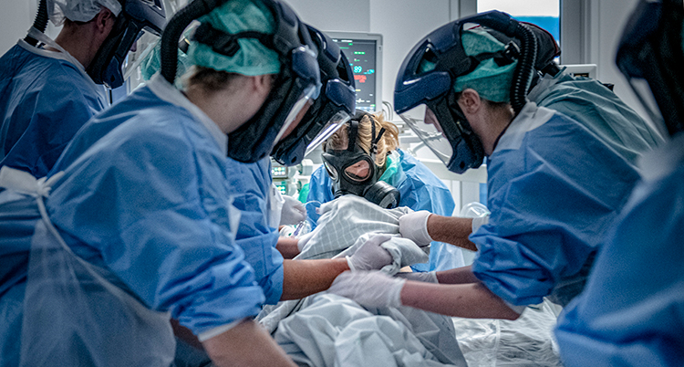 Personalen har skyddskläder. De har handskar och masker för ansiktet. En patient som inte syns ligger ner. Alla står böjda över patienten.