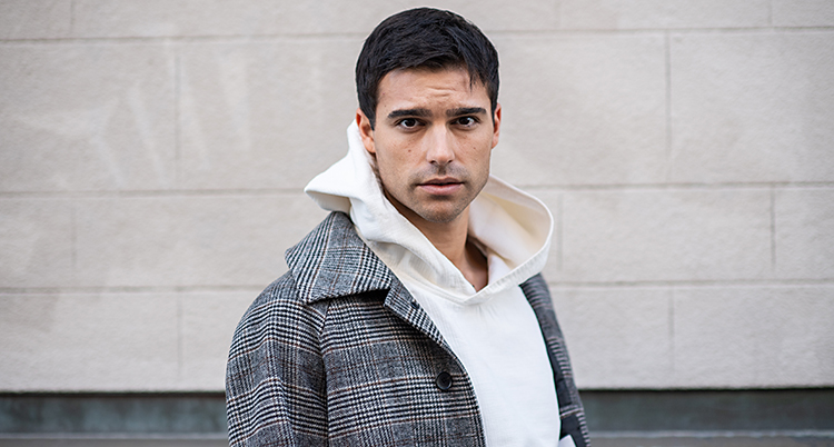 Eric Saade tittar in i kameran. Han har kort mörkt hår. Han har en vit tröja med huva och en grå jacka som är rutig. Han står framför en vit vägg.