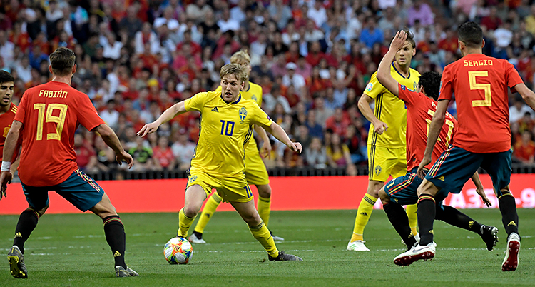 Sveriges herrar spelar en match mot Spanien i fotboll. Den svenska spelaren Emil Forsberg har bollen. Han har gula kläder. Spaniens spelare har röda kläder.