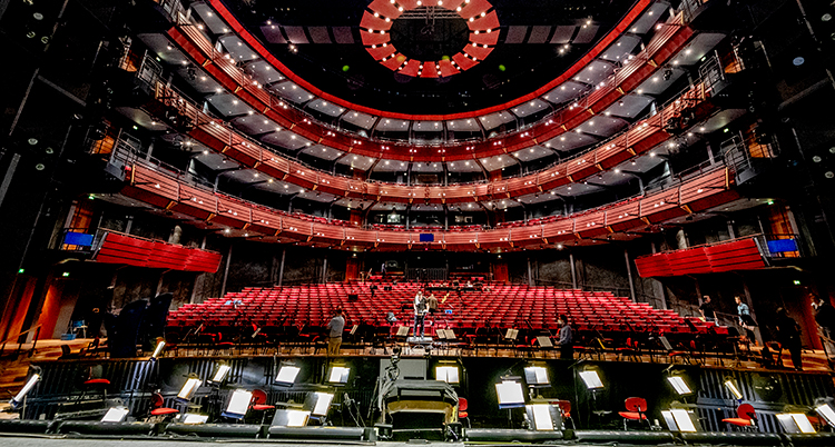 En bild på Göteborgsoperans stora salong tagen från scenen. Det ser maffigt ut med tända lampor i taket och röda sammetsklädda stolar. Salen är tom.