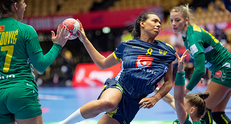 Det är en match i handboll. Jamina Roberts från Sverige försöker skjuta ett skott. Hon har blå kläder. Det andra laget har gröna kläder.