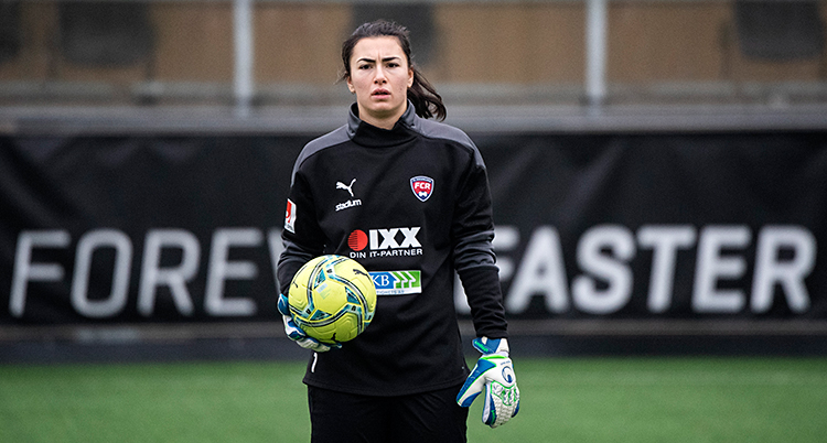 Hon står på en fotbollsplan. Hon har handskar på sig. Hon håller i en fotboll.