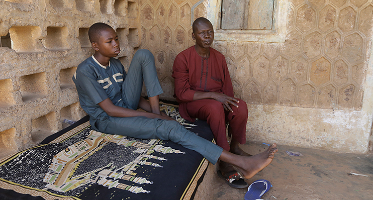 Bilden är tagen i landet Nigeria. En 17-årig pojke sitter på en säng. Han har en blå tröja och blå byxor. Bredvid sitter hans pappa som har röda kläder.