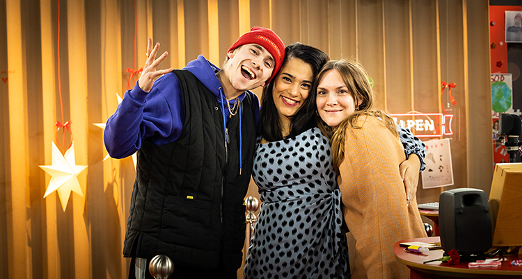 De står i en radiostudio. Felix står till vänster, Farah i mitten och Brita till höger. De är glada och tittar in i kameran.