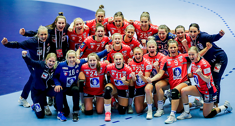 Bilden är tagen inomhus i en sporthall. Vi ser Norges lag i handboll för damer. Alla är samlade på en bild. De jublar och är glada. De har röda kläder.