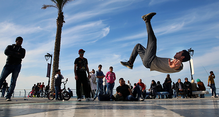 En man dansar breakdance och hoppar i luften med ett ben mot himlen. Han dansar utomhus och bakom honom syns många människor som sitter och står och tittar