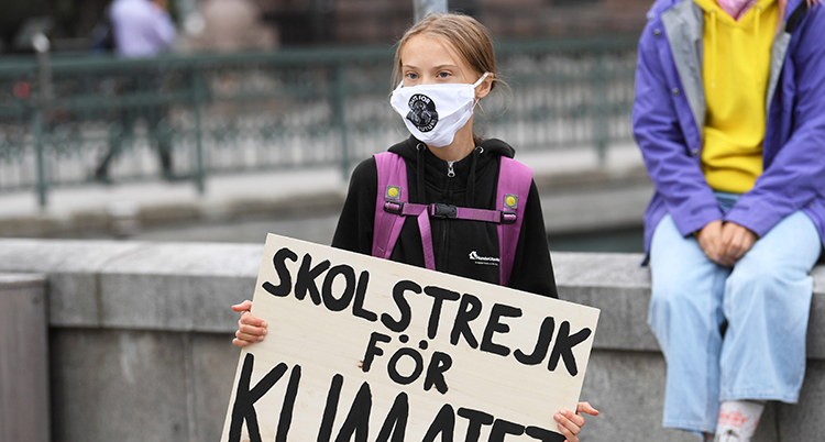Hon håller i en skylt som det står skolstrejk för klimatet på. Hon har munskydd mot viruset corona.
