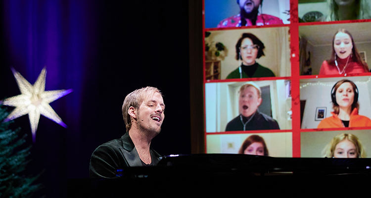 Han sitter vid ett piano. Bakom honom syns en stor skärm med folk som filmat sig själva hemma när de sjunger.